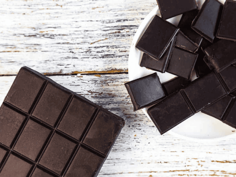 A bar of dark chocolate and several pieces of broken chocolate are displayed on a white wooden surface with some pieces in a white bowl.