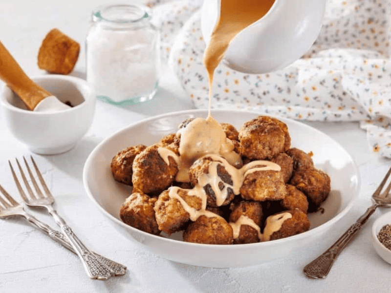 A bowl of meatballs is topped with creamy sauce being poured from a jug, surrounded by forks and seasoning jars on a white table.