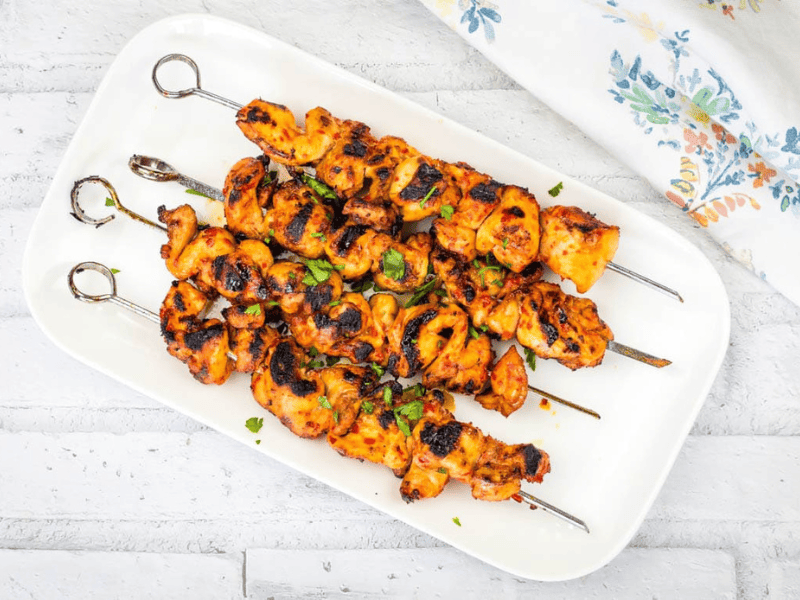 A rectangular white plate with four Sweet & Spicy Grilled Harissa-Honey Chicken Skewers garnished with chopped herbs.