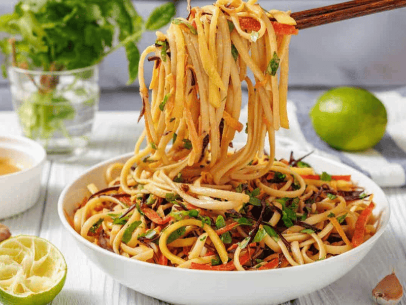 A bowl of Thai Noodle Salad with Mango and Spicy Peanut Dressing is lifted by chopsticks.