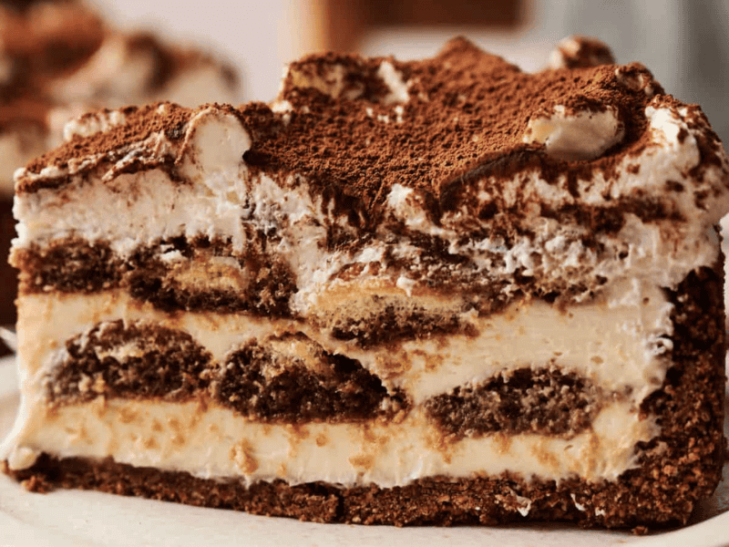 A close-up of a slice of layered tiramisu cake with cream, coffee-soaked sponge, and dusted cocoa powder on top, served on a white plate.