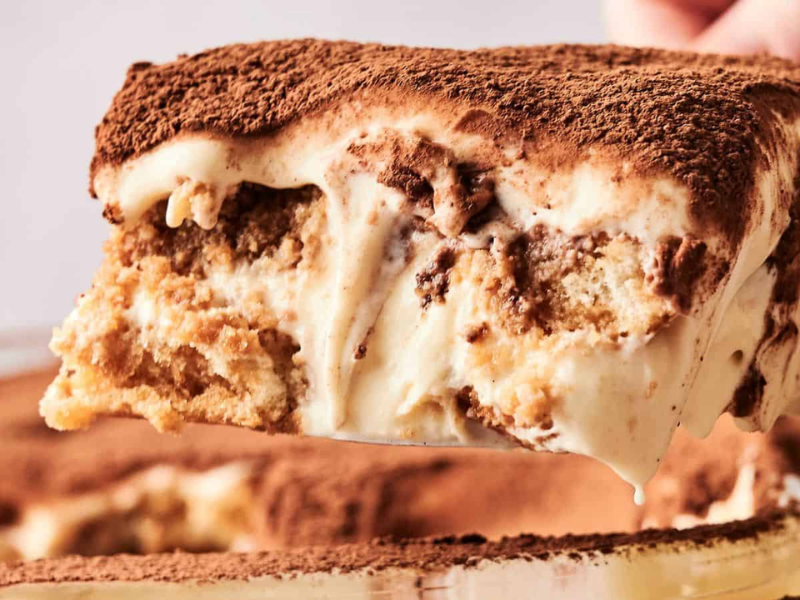 A slice of tiramisu is being lifted from the dish, showcasing layers of coffee-soaked ladyfingers and creamy mascarpone, topped with cocoa powder.