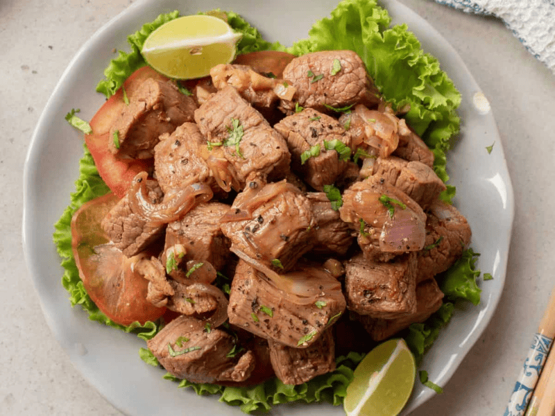 A plate of stir-fried beef cubes with onions, garnished with cilantro, served on lettuce with tomato slices and lime wedges. Chopsticks rest beside the plate.
