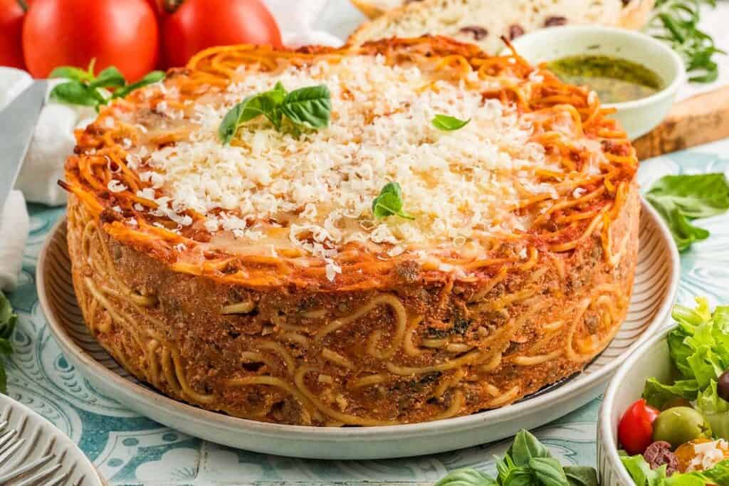 A round baked spaghetti pie makes a comforting pasta dinner, topped with grated cheese and basil, served on a plate with salad, bread, tomatoes, and pesto in the background.