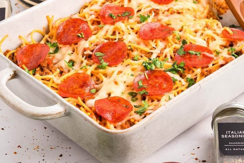 Rectangular baking dish filled with baked spaghetti, melted cheese, pepperoni slices, and chopped parsley—perfect for lovers of Pasta Classics. An Italian seasoning jar sits nearby.