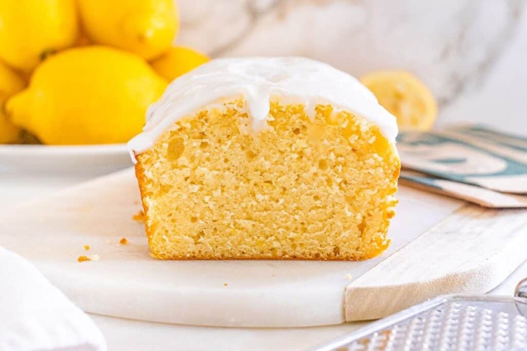 An easy desserts favorite&mdash;a sliced lemon loaf cake with white icing on top sits on a round white cutting board; lemons are visible in the background.