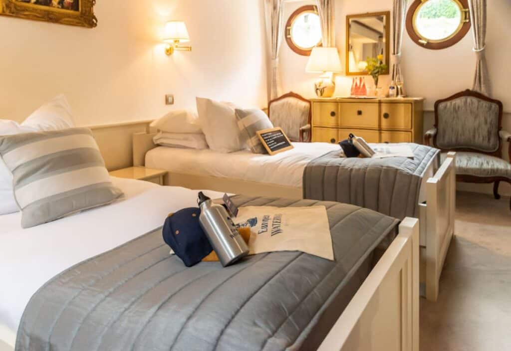 Two single beds with gray bedding in a neatly arranged room aboard a luxury barge cruise, featuring a dresser, armchair, round windows, and soft lighting from wall sconces and a lamp.