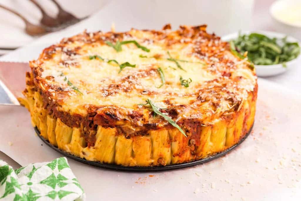 A baked pasta pie with rigatoni noodles standing upright, topped with melted cheese, tomato sauce, and fresh herbs on a white surface&mdash;a perfect pasta dinner for any occasion.