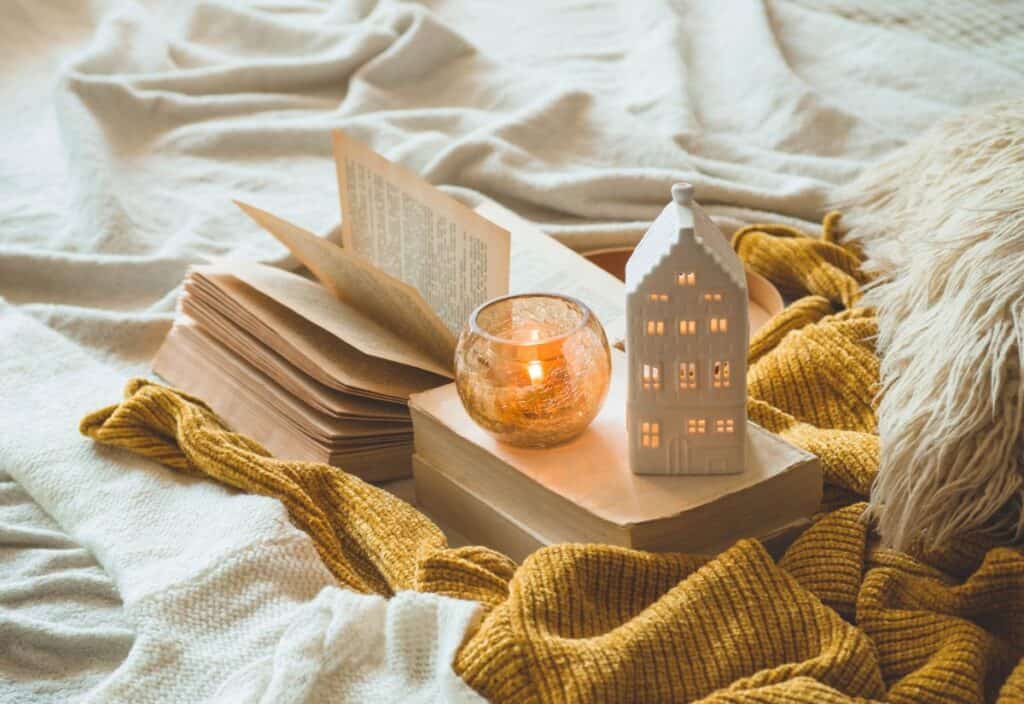 An open book, a lit candle in a glass holder, and a ceramic house sit on a blanket&mdash;cozy up with the yellow knitted throw and pillow in the background.