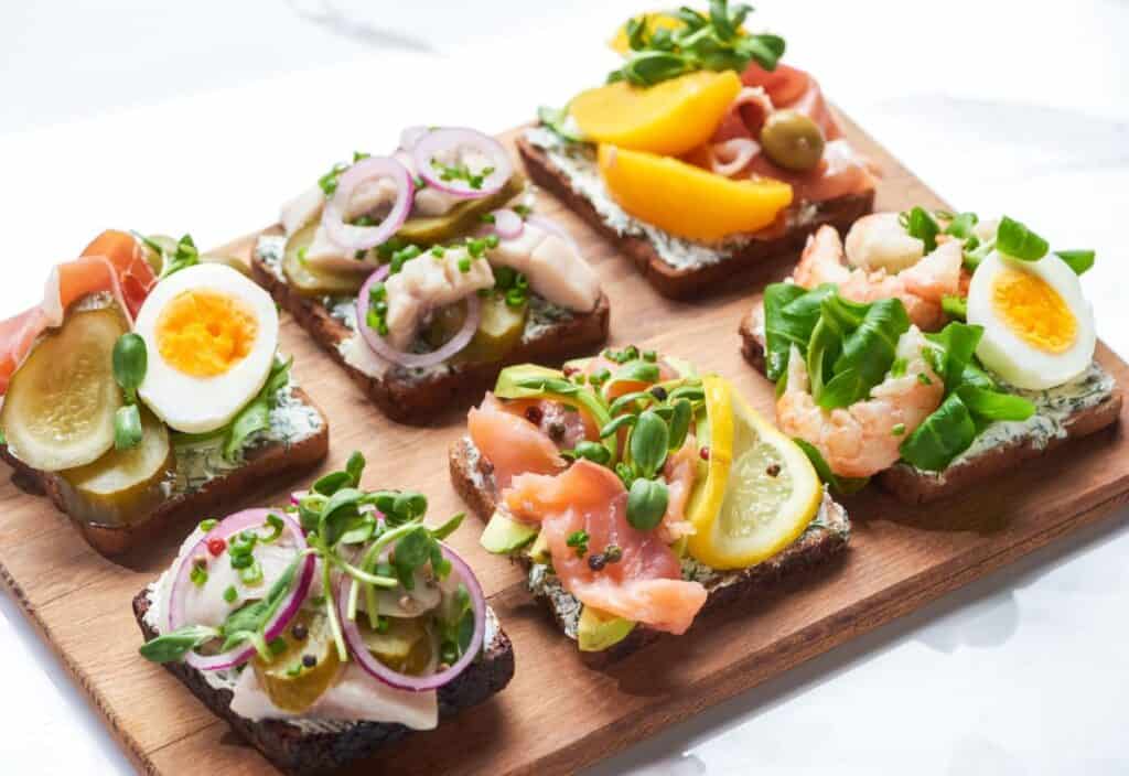 Six open-faced sandwiches with various toppings, including sliced eggs, smoked salmon, shrimp, pickles, red onions, greens, and peach slices, arranged on a wooden serving board.