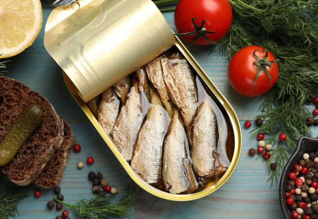 An opened tin of tinned seafood in oil is surrounded by tomatoes, dill, black bread with a pickle, lemon, and assorted peppercorns on a wooden surface.