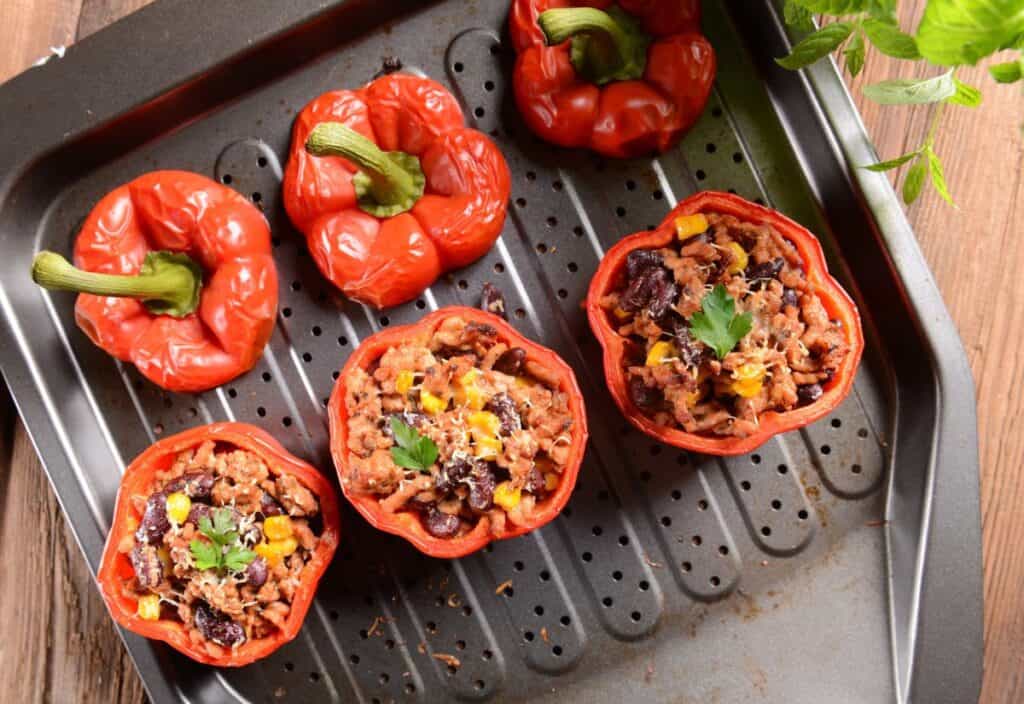 Three red bell peppers stuffed with a mixture of rice, beans, corn, and herbs on a roasting tray, with three unstuffed pepper tops beside them.