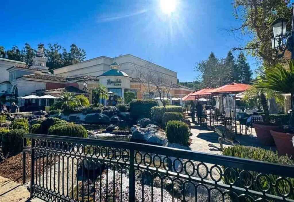 Outdoor dining area with tables and umbrellas near a garden and small stream, under a clear blue sky with the sun shining brightly.
