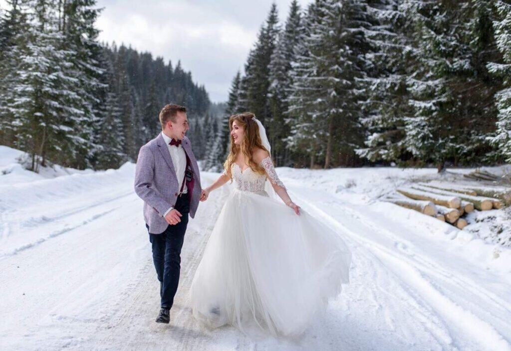 A bride and groom walk hand in hand on a snow-covered road in a forest, surrounded by pine trees and dressed in wedding attire—capturing the magic of winter weddings.