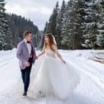 A bride and groom walk hand in hand on a snow-covered road in a forest, surrounded by pine trees and dressed in wedding attire&mdash;capturing the magic of winter weddings.