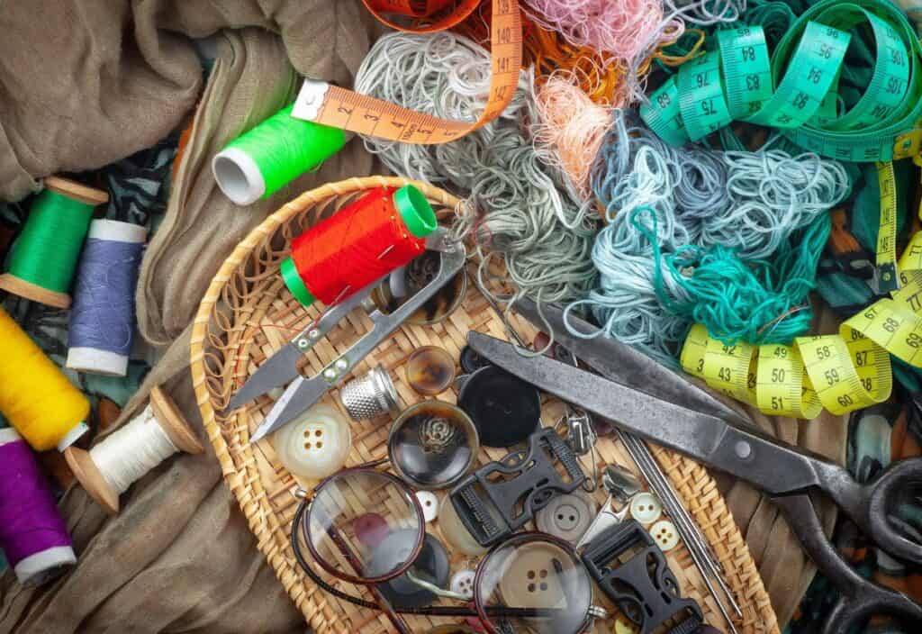 A basket with assorted sewing supplies, including thread spools, buttons, needles, and scissors&mdash;perfect for making do&mdash;sits among colorful yarn and fabric, with measuring tapes and eyeglasses completing the scene.