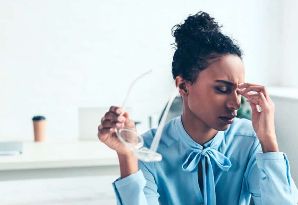 Woman in a blue blouse sitting at a desk, holding her glasses in one hand and pinching the bridge of her nose with a pained expression.