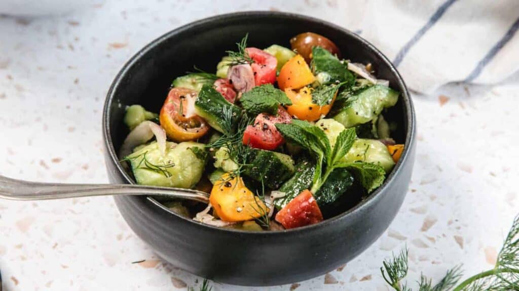 A black bowl filled with a fresh salad of sliced cucumbers, cherry tomatoes, red onions, herbs, and black pepper, with a spoon on the side.