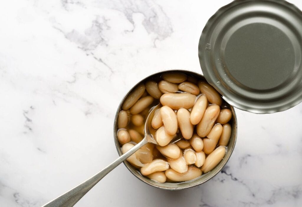 An opened can of white beans in liquid, with a spoon inside, sits on a white marble surface next to the removed lid.