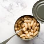 An opened can of white beans in liquid, with a spoon inside, sits on a white marble surface next to the removed lid.