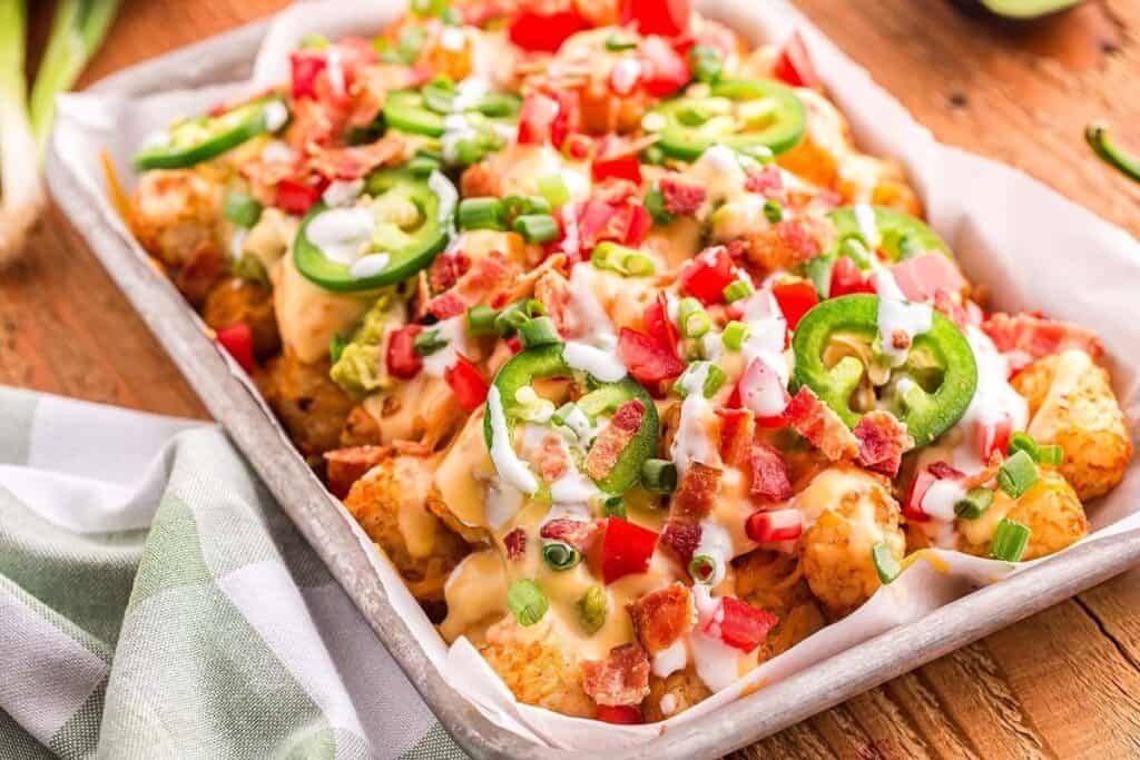 A tray of loaded tater tots topped with melted cheese, diced tomatoes, jalape&ntilde;o slices, bacon bits, green onions, and drizzled with white sauce&mdash;perfect Game Day Snacks on a wooden table.