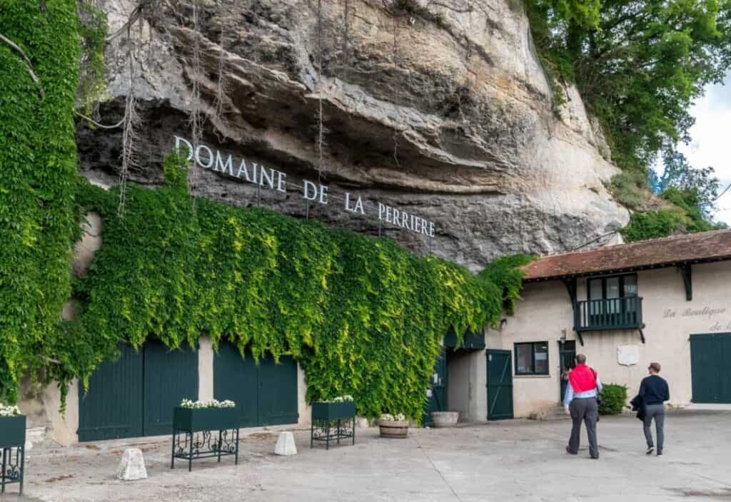Two people walk toward a stone building built into a rocky hillside, with "Domaine de la Perri&egrave;re" above green doors and ivy covering part of the facade&mdash;a charming stop on a luxury barge cruise through scenic countryside.