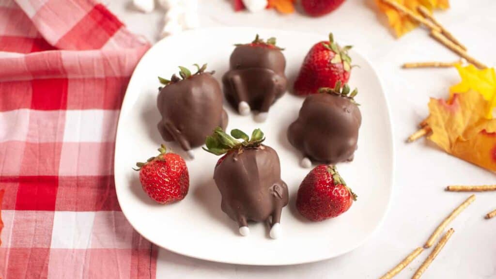 A plate with strawberries dipped in chocolate, some decorated to look like animal bottoms with candy feet, alongside plain strawberries, on a white surface with a red checked cloth and autumn leaves.