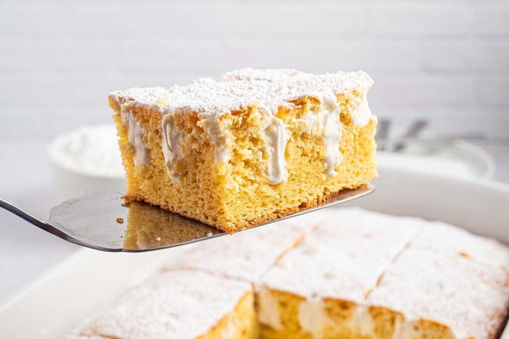 A piece of yellow sponge cake with white icing and powdered sugar is being lifted from a pan containing similar pieces&mdash;perfect for those seeking easy desserts to make at home.