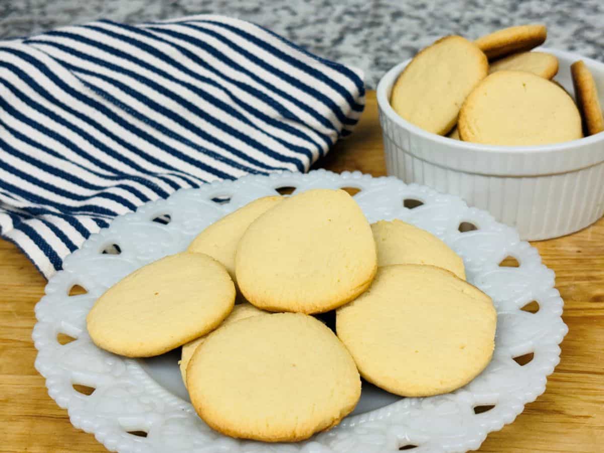A white plate with round, golden-brown cookies sits on a wooden surface next to a bowl of cookies and a blue-and-white striped cloth.