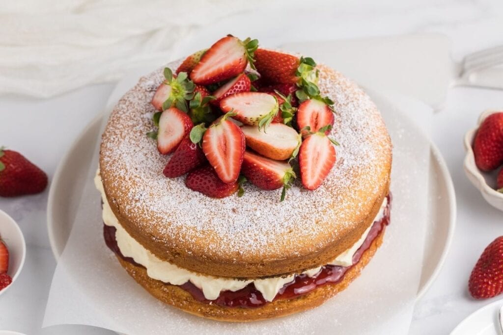 A sponge cake inspired by Bridgerton recipes, filled with cream and strawberry jam, is topped with fresh halved strawberries and a dusting of powdered sugar, elegantly presented on a white plate.