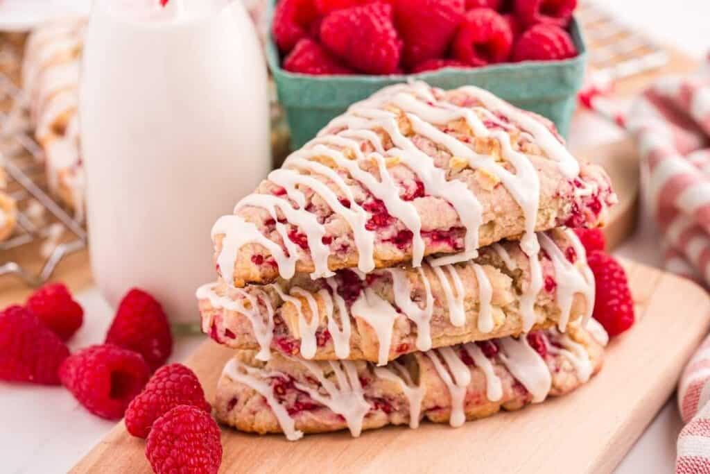 Three raspberry scones with white icing are stacked on a wooden board, evoking classic Bridgerton recipes, with fresh raspberries and a bottle of milk in the background.