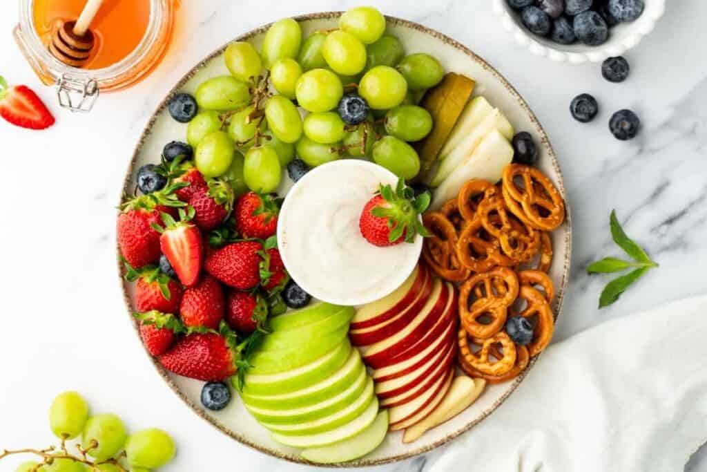 A round plate with sliced green and red apples, strawberries, blueberries, grapes, pear slices, pretzels, and a bowl of dip in the center&mdash;this elegant snack platter is perfect for serving at a Bridgerton recipes-inspired gathering.