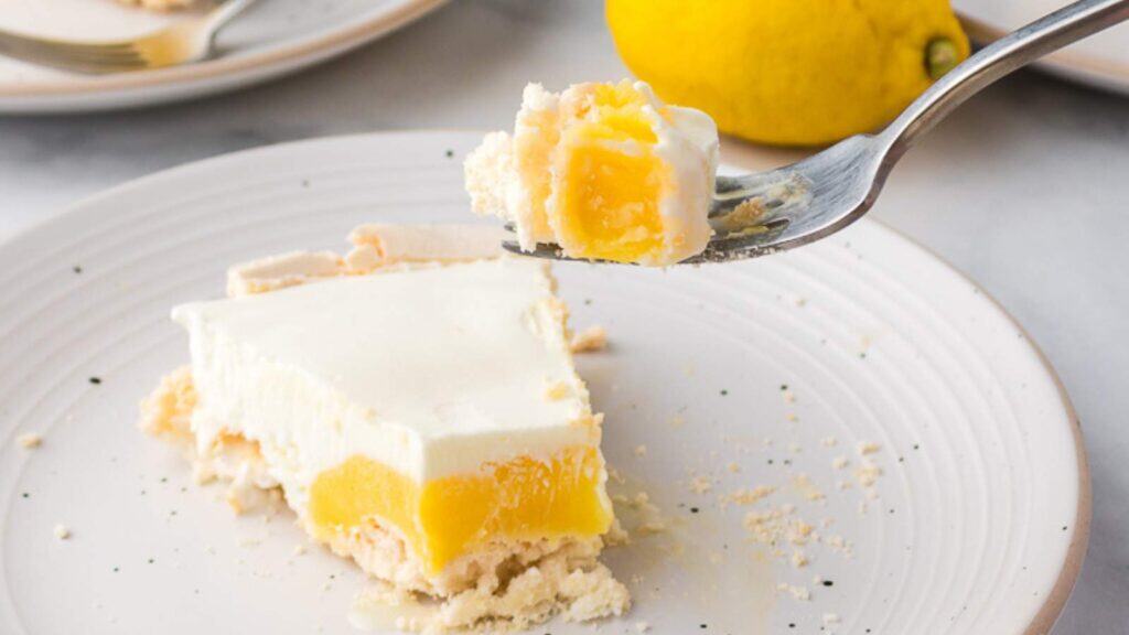A fork holds a bite of creamy lemon dessert with a yellow fruit layer and crumbly crust; a slice and a whole lemon are on the plate in the background.