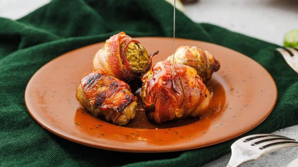 Three bacon-wrapped cabbage rolls on a brown plate, with a drizzle of sauce being poured over them, set on a green cloth.