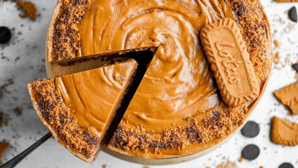 A cheesecake topped with smooth caramel spread, a whole Lotus biscuit, and a crumb border, with one slice being lifted out.