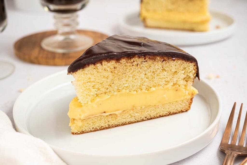 A slice of Boston cream pie with yellow custard filling and chocolate glaze on top, served on a white plate with a fork beside it&mdash;one of the classic easy desserts to enjoy anytime.