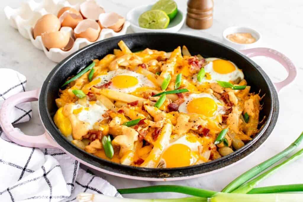 A skillet filled with baked fries, melted cheese, eggs, bacon, and green onions sits on a countertop&mdash;a delicious option for those seeking tasty breakfast ideas&mdash;next to eggshells, limes, a pepper grinder, and a kitchen towel.