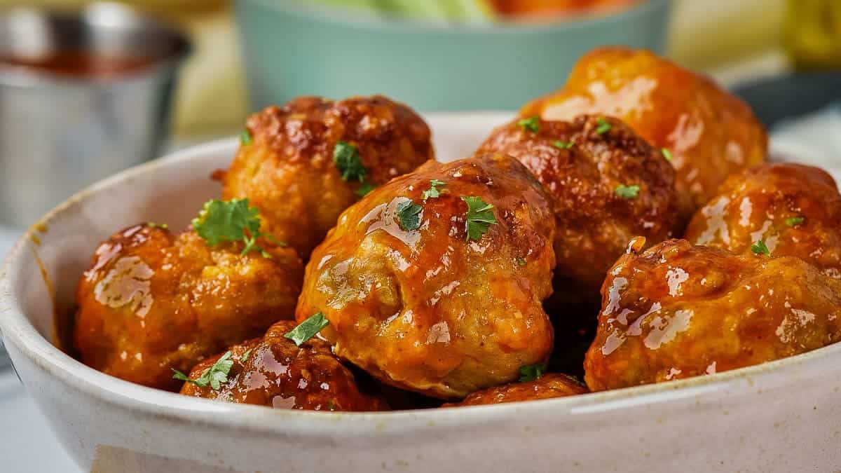 A close-up of a white dish filled with glazed meatballs garnished with chopped herbs.