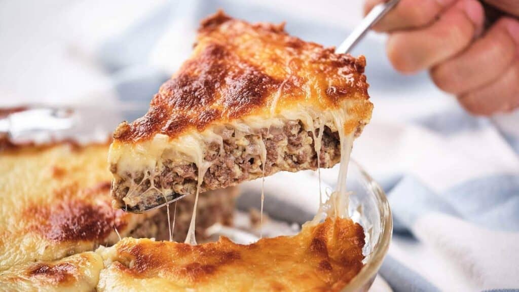 A hand lifts a cheesy, golden-brown slice of baked meat and cheese casserole from a glass dish.