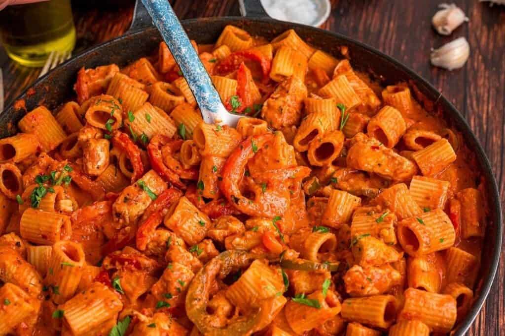 A skillet filled with rigatoni Pasta Classics, sliced bell peppers, and chicken pieces in a creamy tomato sauce, garnished with chopped herbs.