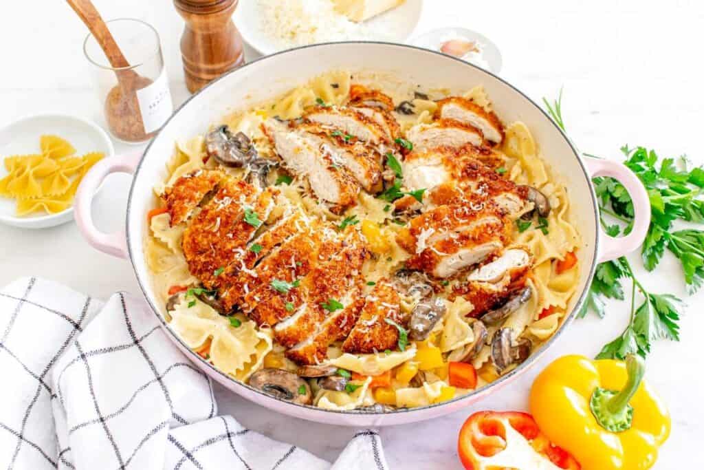 This low effort dinner features breaded chicken slices over farfalle pasta with mushrooms, bell peppers, and creamy sauce, garnished with parsley and grated cheese—all prepared in a single skillet.