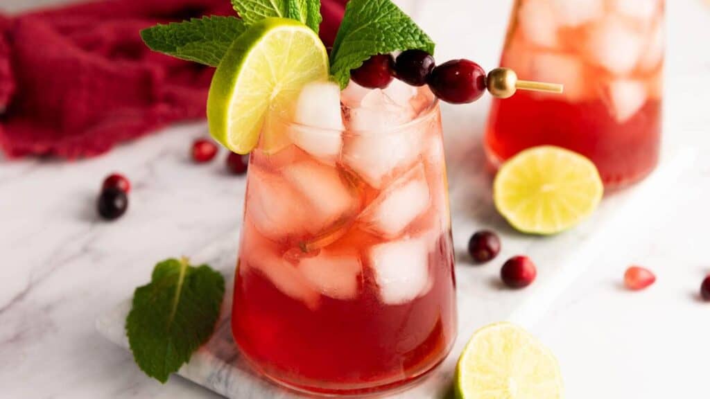 A glass of red iced drink garnished with a lime slice, mint leaves, and cranberries on a skewer, on a marble surface with lime, cranberries, and mint nearby.