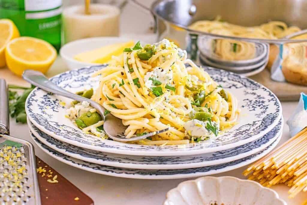 A plate of spaghetti with green vegetables and cheese, garnished with herbs, sits on stacked plates with a spoon and fork; Pasta Classics like this shine among lemons and kitchen items in the background.
