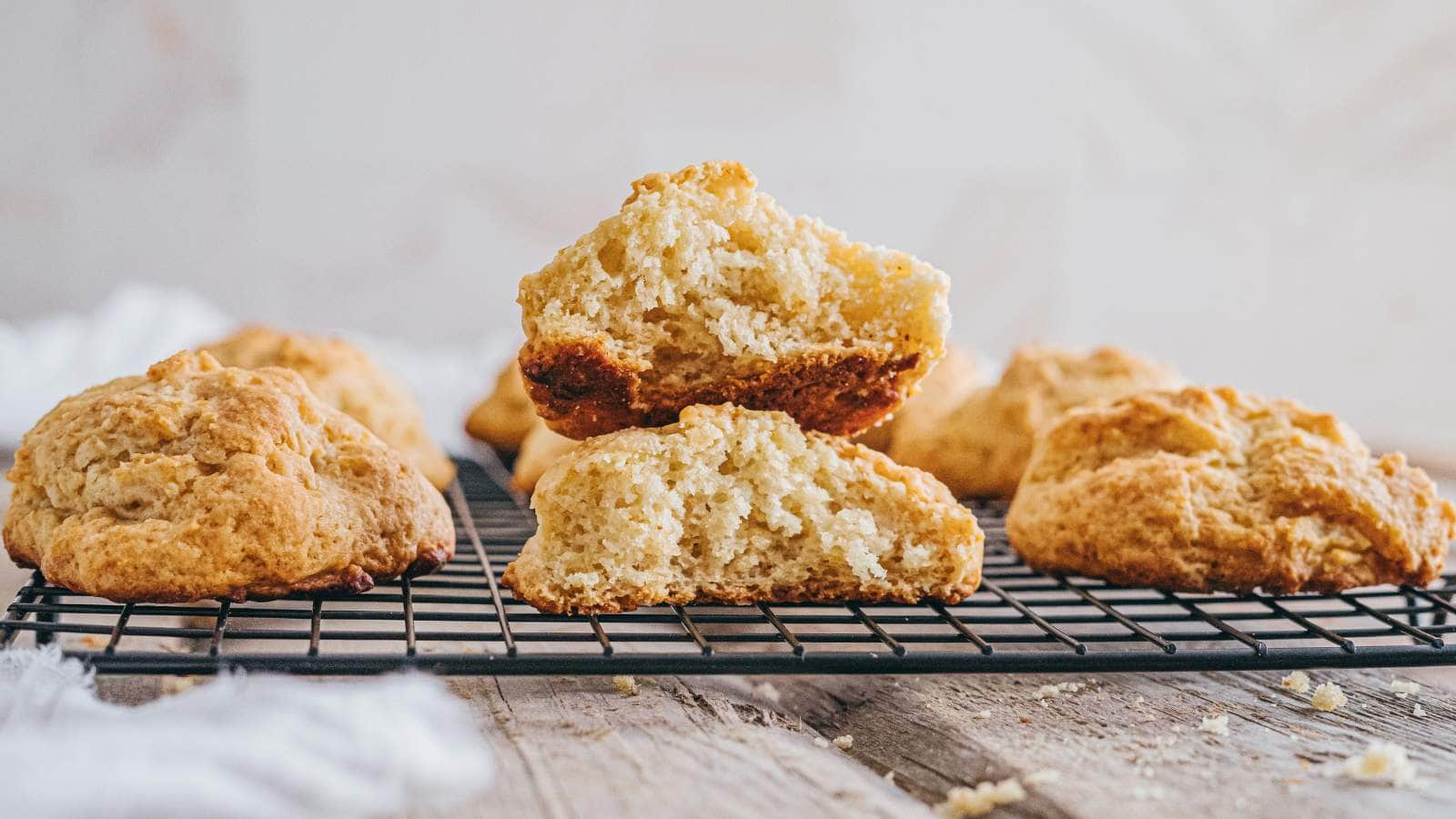 Golden brown drop biscuits cooling on a wire rack, with one biscuit split open to show its soft, fluffy interior.