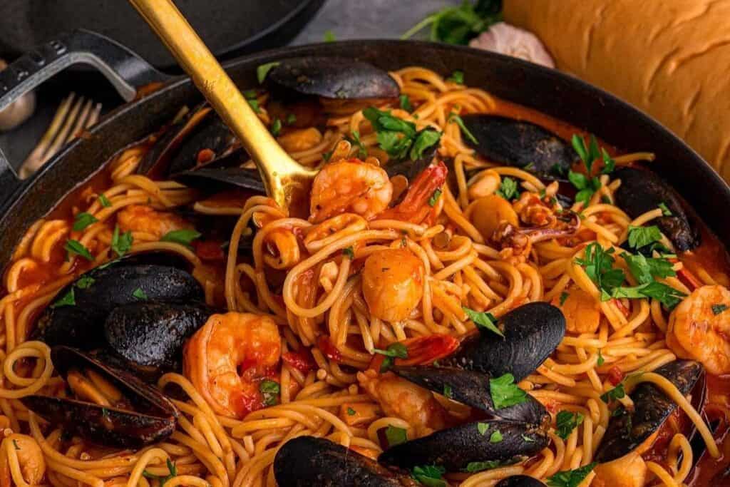 Spaghetti with shrimp, mussels, and tomato sauce—one of the Pasta Classics—garnished with parsley in a skillet, accompanied by a serving spoon and bread in the background.