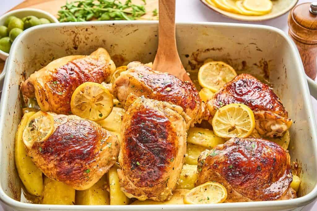 Roasted chicken thighs with browned skin, sliced lemons, and potato wedges in a baking dish—a perfect option for low effort dinners.