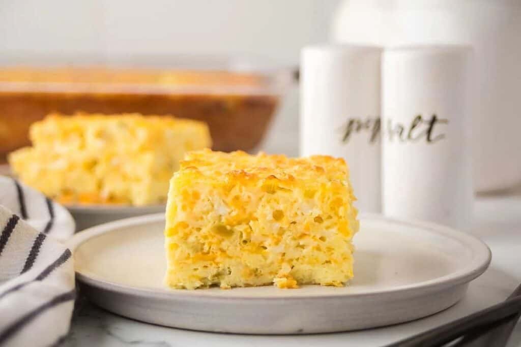 A square slice of baked macaroni and cheese sits on a round plate, perfect for those seeking tasty breakfast ideas, with a baking dish and salt and pepper shakers in the background.
