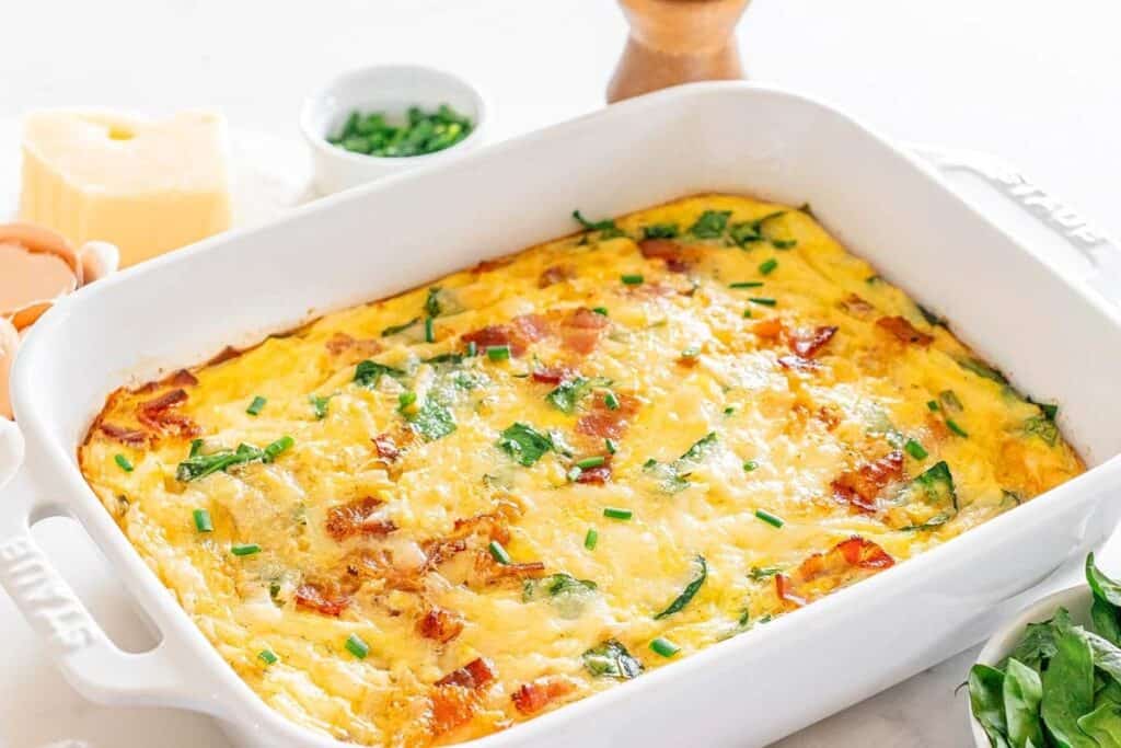 A baked egg casserole with spinach, cheese, and bacon in a white rectangular dish&mdash;surrounded by eggs, cheese, and chives&mdash;makes for one of the tastiest breakfast ideas to start your day right.