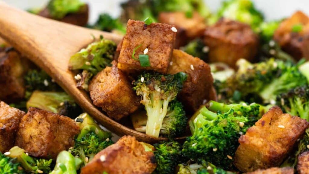 A wooden spoon lifts pieces of roasted tofu and broccoli, garnished with sesame seeds and chopped green onions.