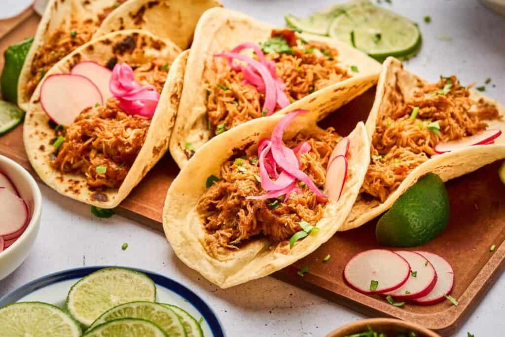 Four shredded chicken tacos topped with pickled onions and sliced radishes on a wooden board, with lime wedges and more radish slices nearby—a perfect choice for low effort dinners.